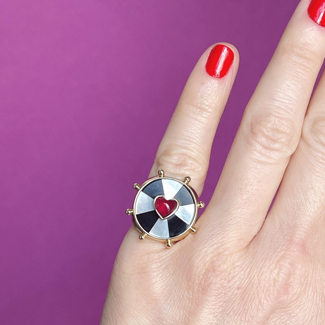 Hand against a purple background showing the yellow gold ring with a black and white bullseye pattern and pink heart set in the center