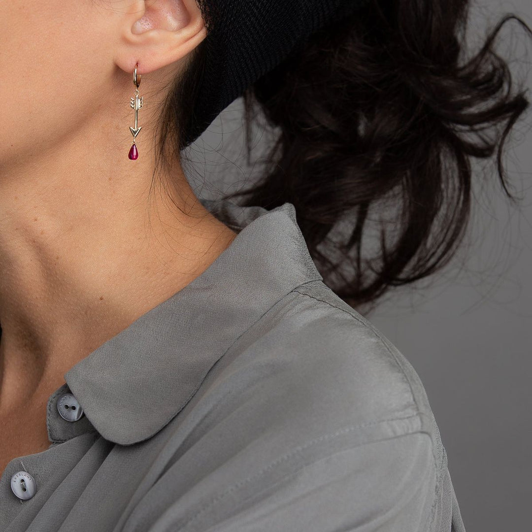 Woman in grey wearing a yellow gold arrow earring on a closed hoop with a pink ruby dripping from the arrow's tip