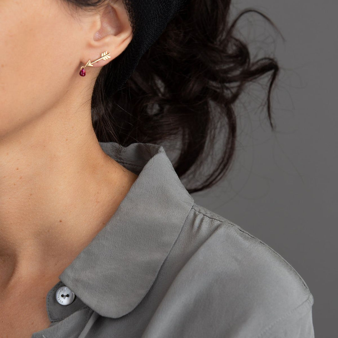 model in grey wearing a single yellow gold arrow stud with pink ruby droplets dangling from the arrows's tip