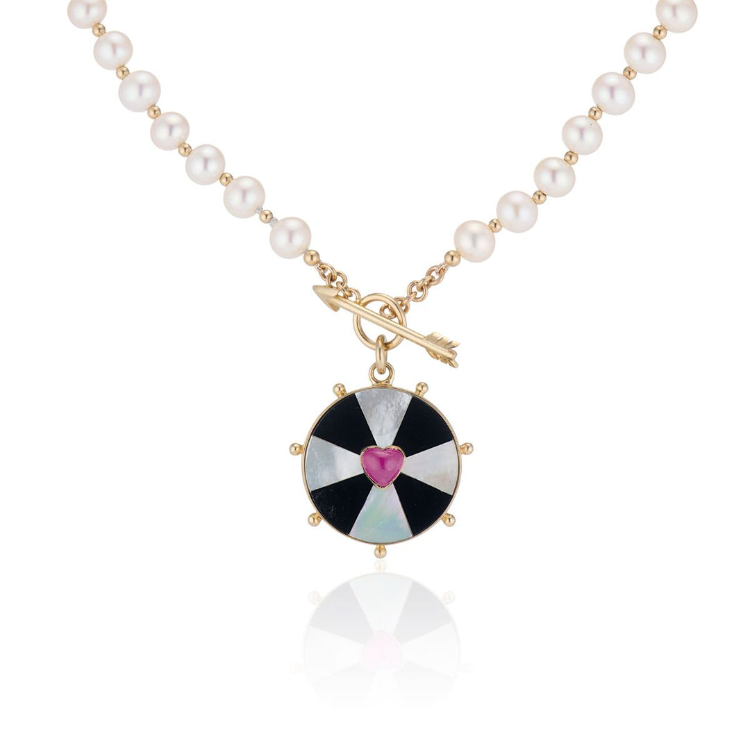 black and white alternating bullseye circle pendant with pink heart in center, hanging on a pearl and gold chain necklace with arrow clasp in front