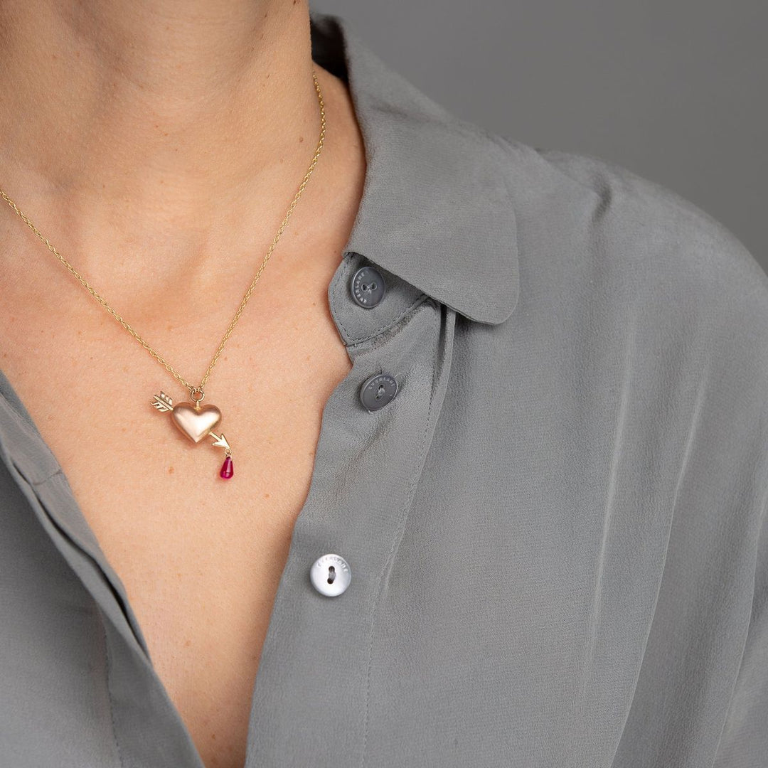 woman in grey blouse modeling small rose gold puffed heart pendant on yellow gold chain with yellow gold arrow piercing the heart with a single ruby dripping from tip of arrow