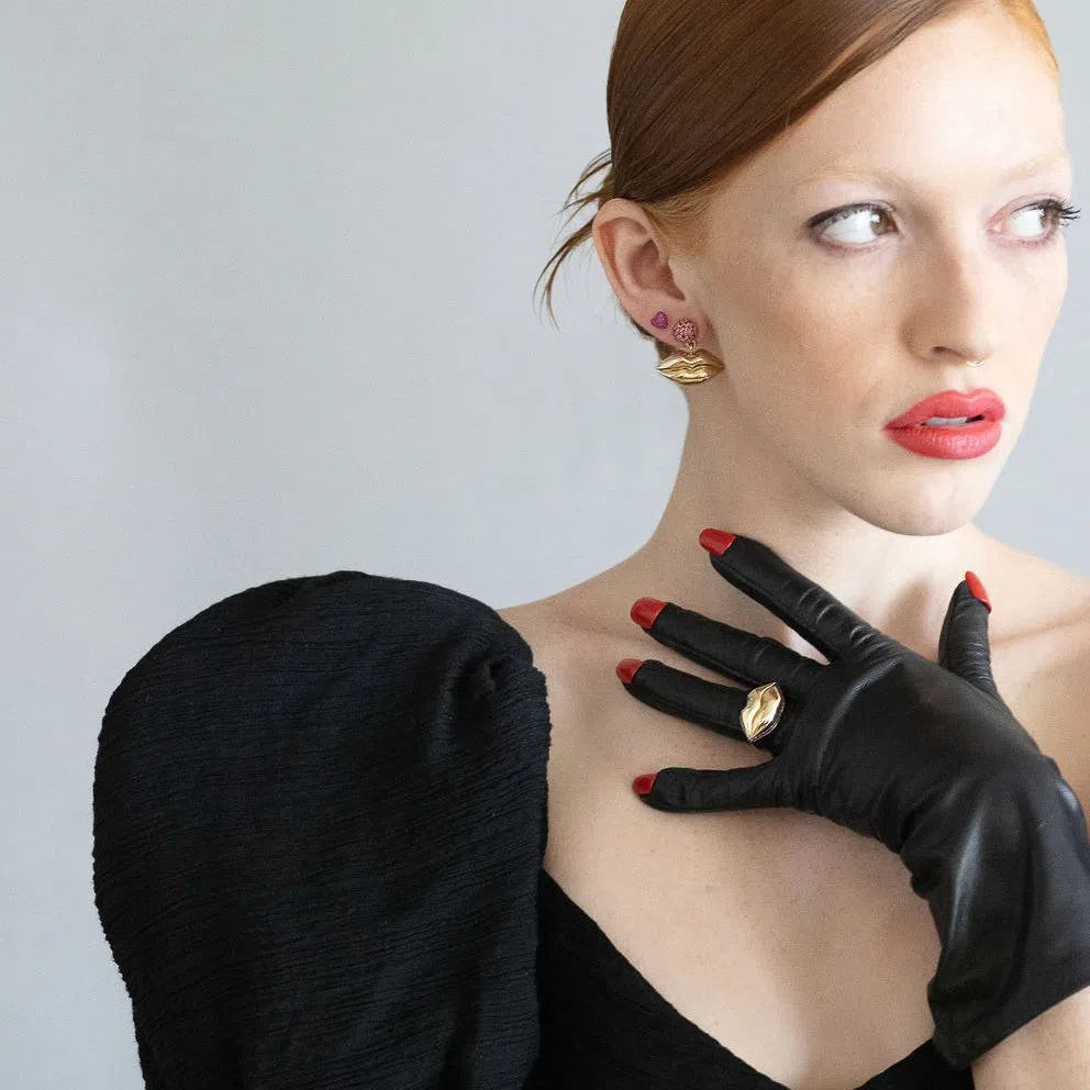 red head model wearing gold lips earrings and gold lip ring on her hand over black gloves next to her mouth on a grey background