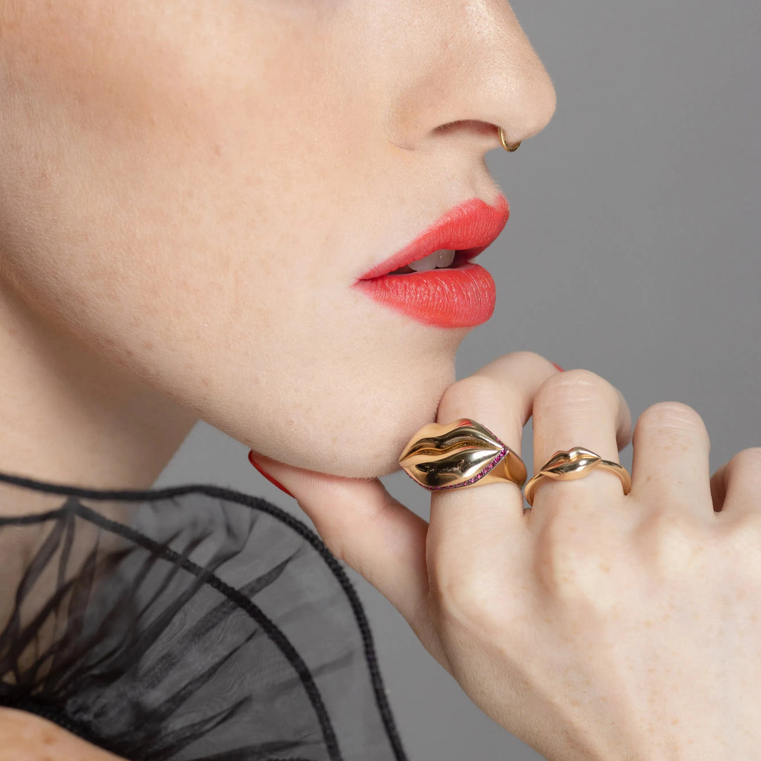 red head model wearing gold lip ring on her hand next to her mouth on a grey background