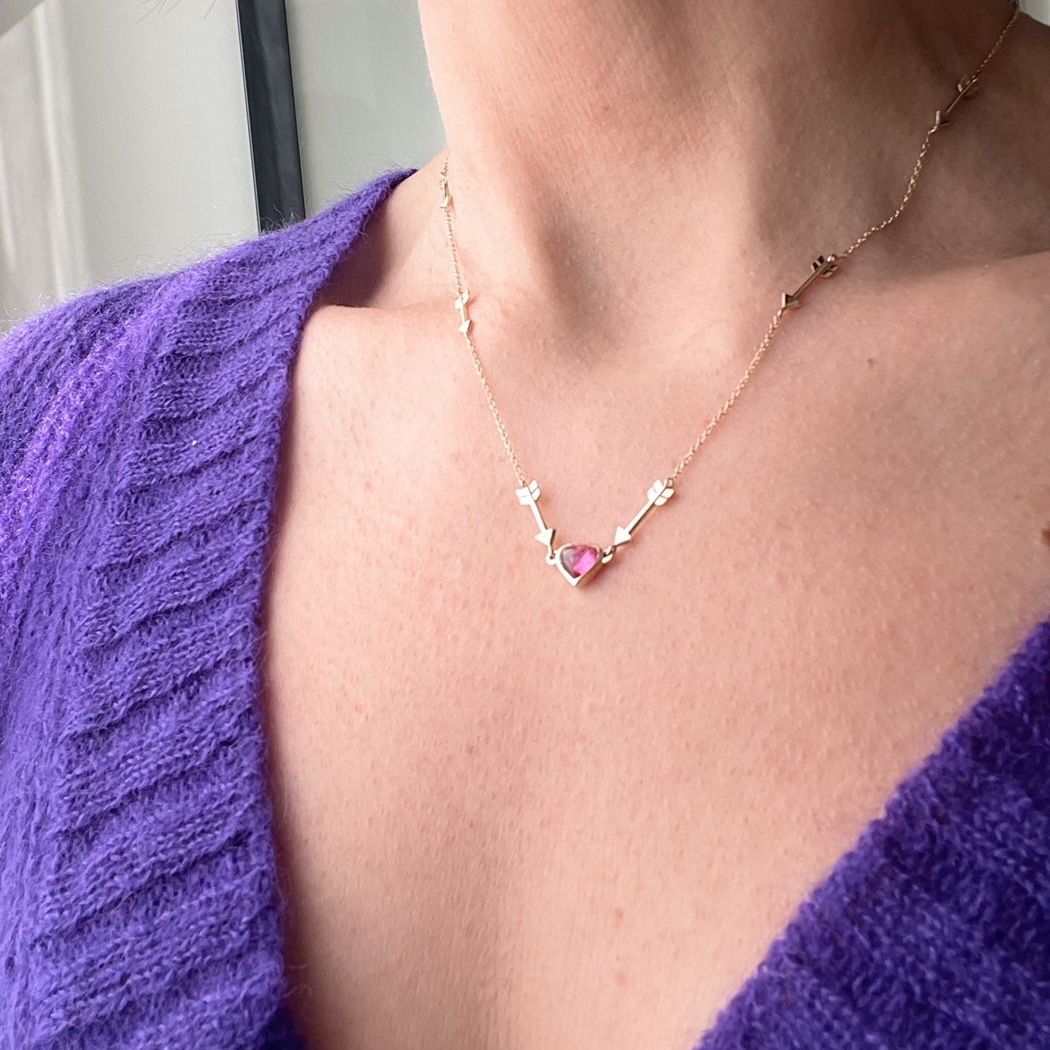 Close up on a neck of a yellow gold chain with small arrow spaced intermittently pointing to the pink heart at the center