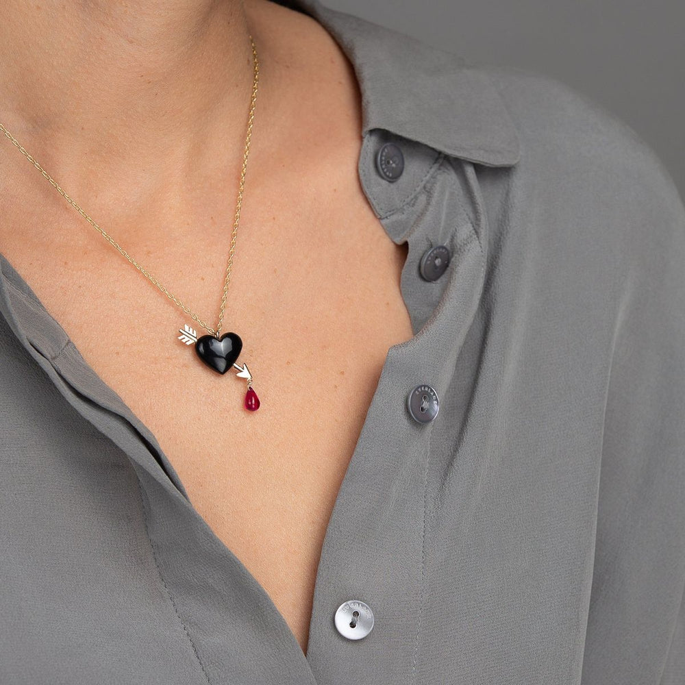 woman in grey modeling small Smooth jet black onyx heart is pierced with a golden arrow, dripping a single droplet of ruby blood on gold chain necklace