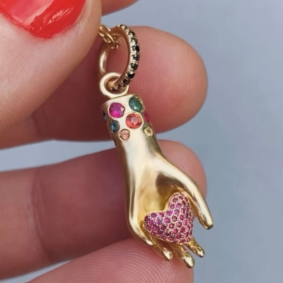 close up shot of small hand charm in yellow gold holding puffed heart encrusted with rubies, gemstones set all around wrist, and black diamonds set all on bail. on delicate yellow gold chain necklace