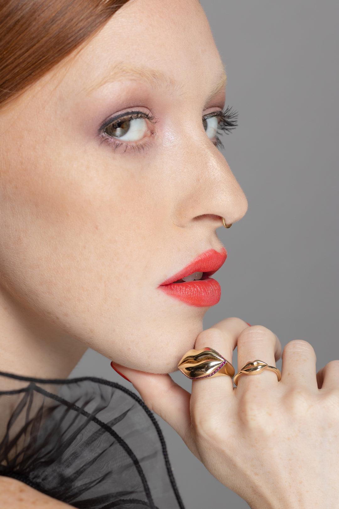 red head model wearing gold lip ring on her hand next to her mouth on a grey background