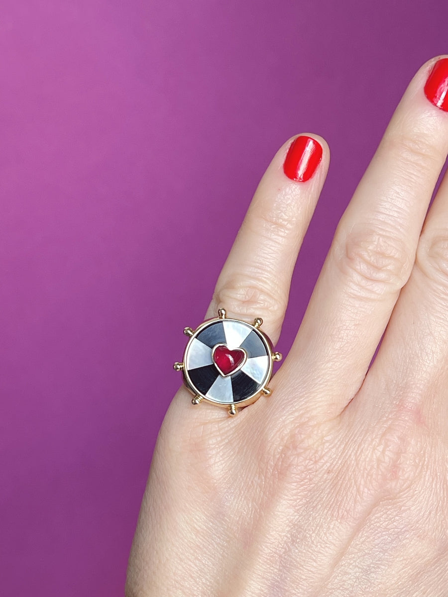 Hand against a purple background showing the yellow gold ring with a black and white bullseye pattern and pink heart set in the center
