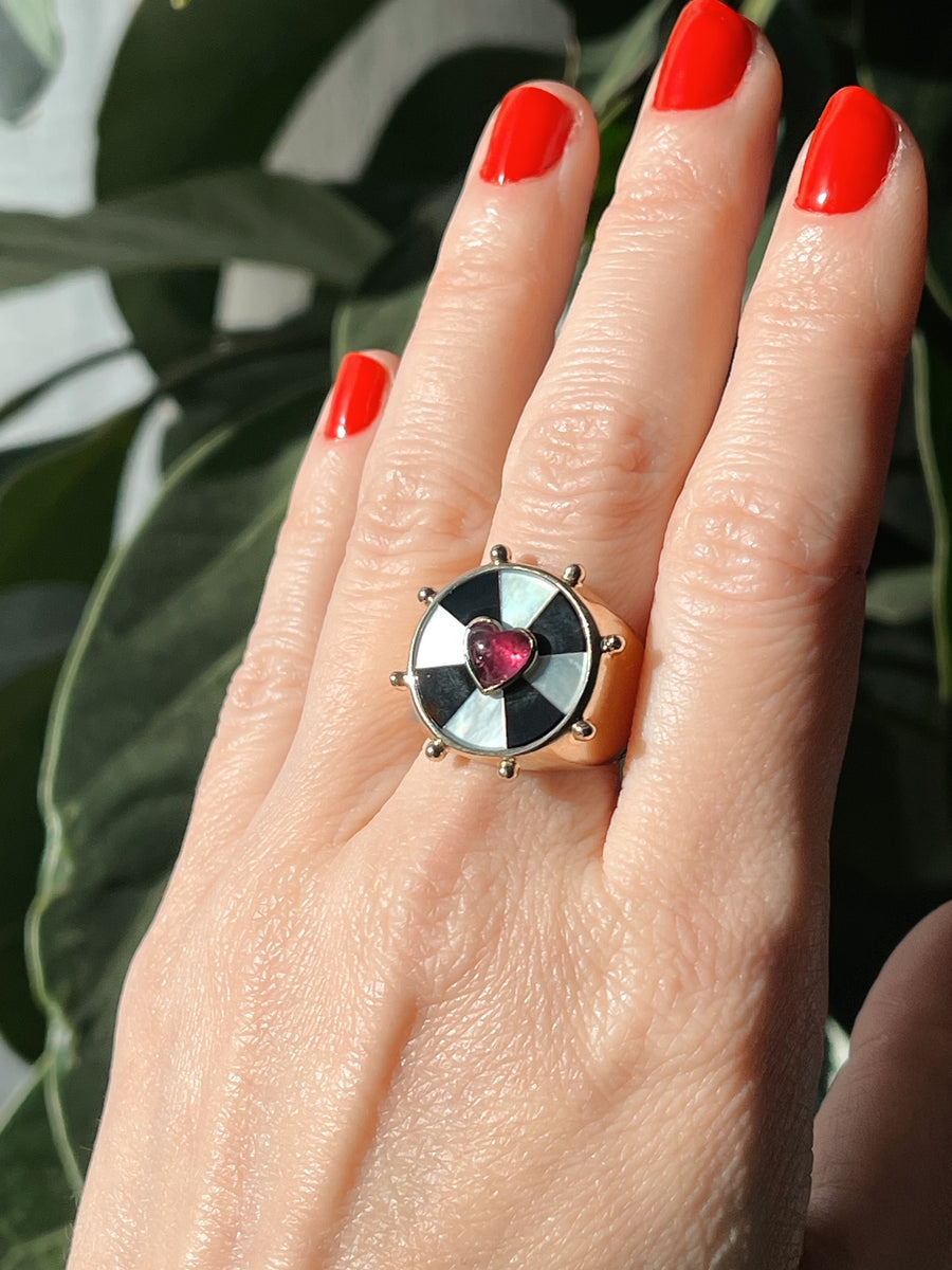 Hand with red nail against greenery showing the yellow gold ring with a black and white bullseye pattern and a pink heart set in the center