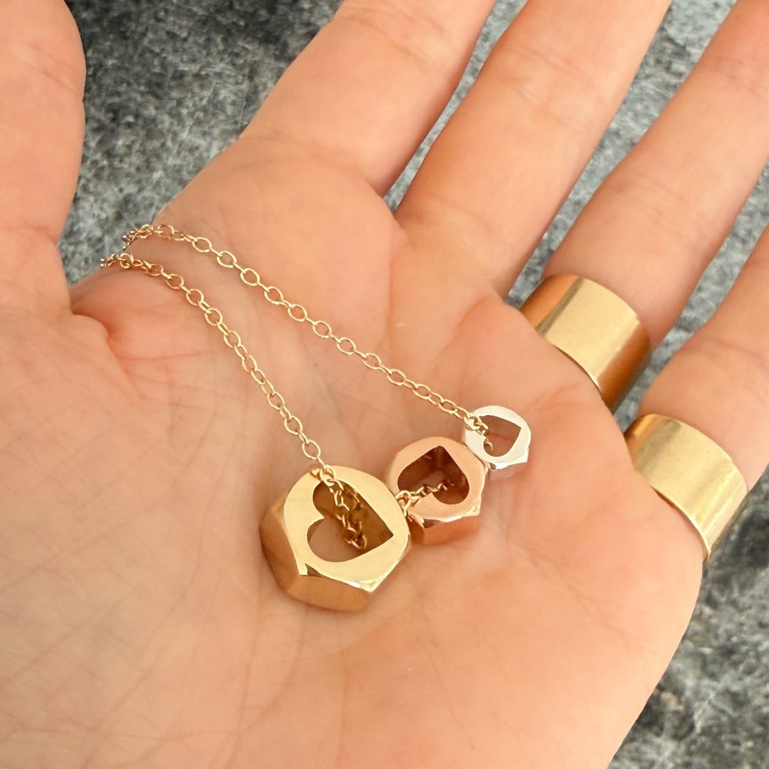 Three hex nut charms strung on gold chain in 3 different sizes in yellow and rose gold, and silver laying flat in a hand wearing 2 gold bands. 