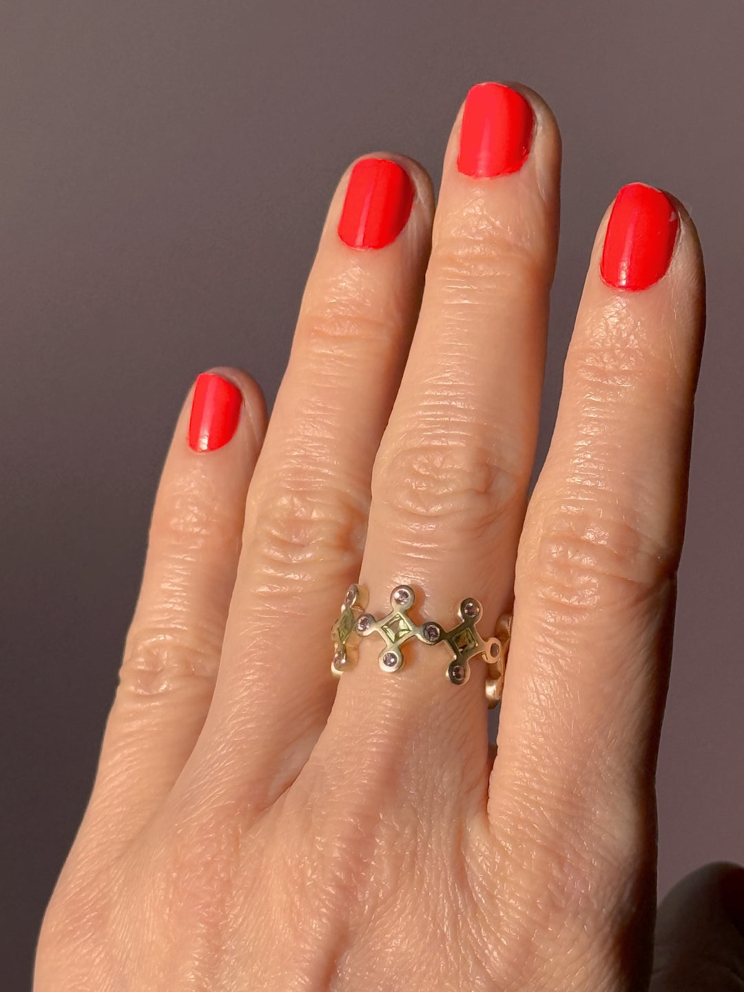 Yellow gold ring with pink and green gemstones on a hand with red nails on a neutral background