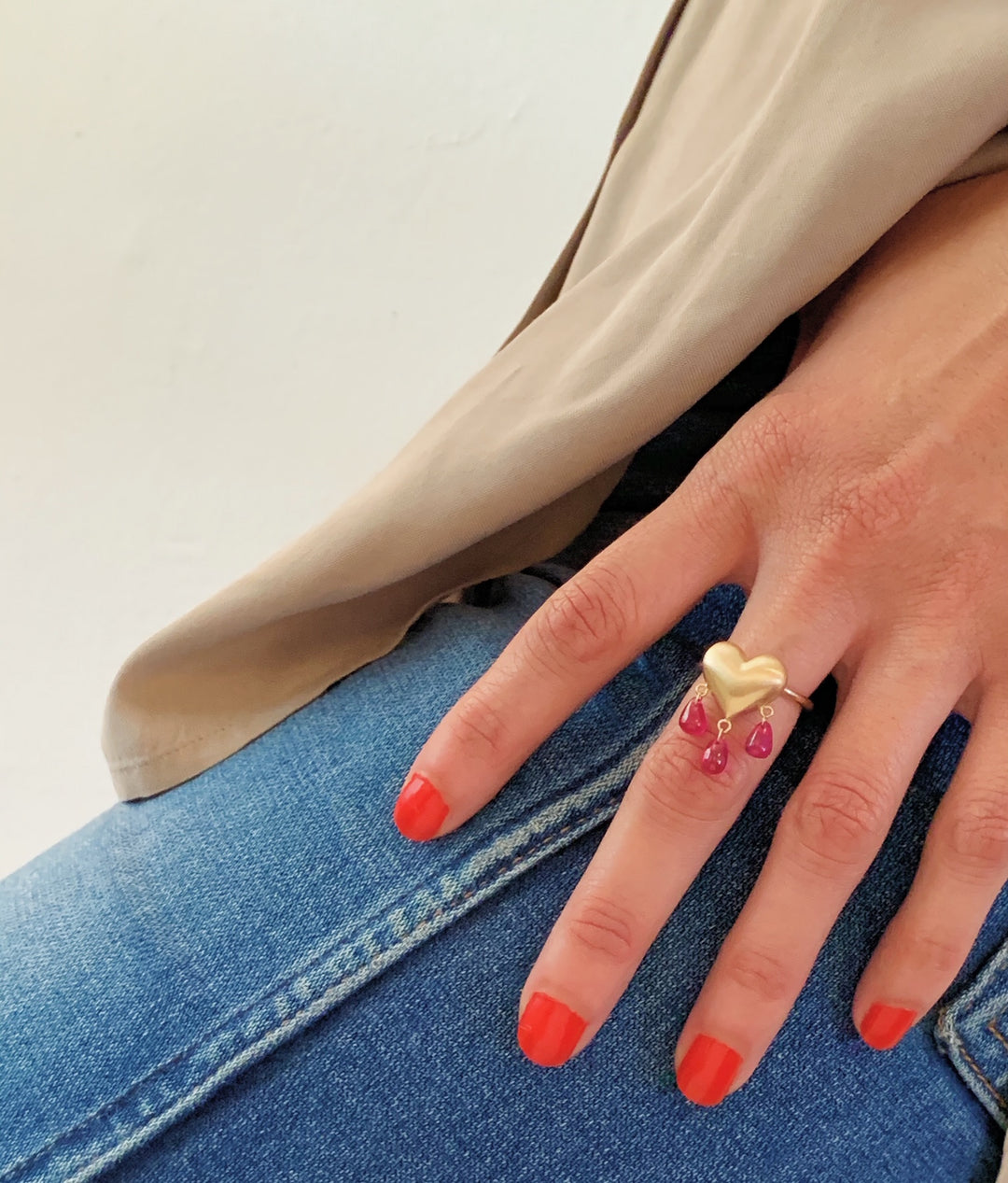 Hand wearing a gold heart ring with red ruby droplets hanging below against denim fabric.