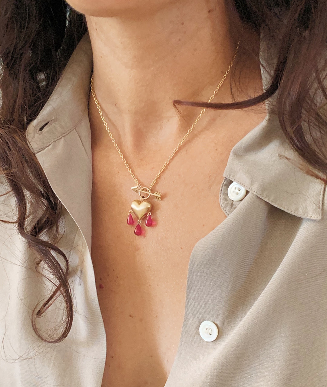 Close up of a woman with brown hair wearing a gold heart pendant necklace with red ruby droplets and an arrow front toggle clasp.