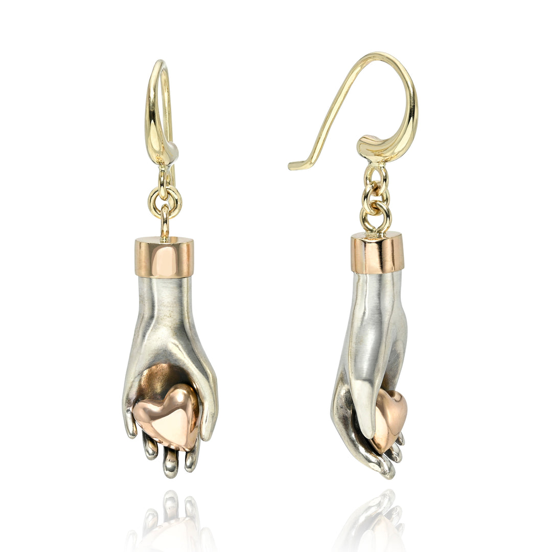 Pair of silver and gold hand earrings with holding rose gold hearts on a white background