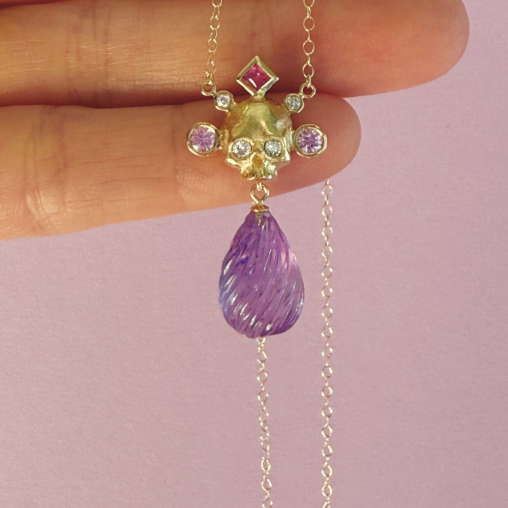 A gold skull pendant necklace with a purple teardrop gemstone and pink and purple stones, held between fingers against a soft pink background.
