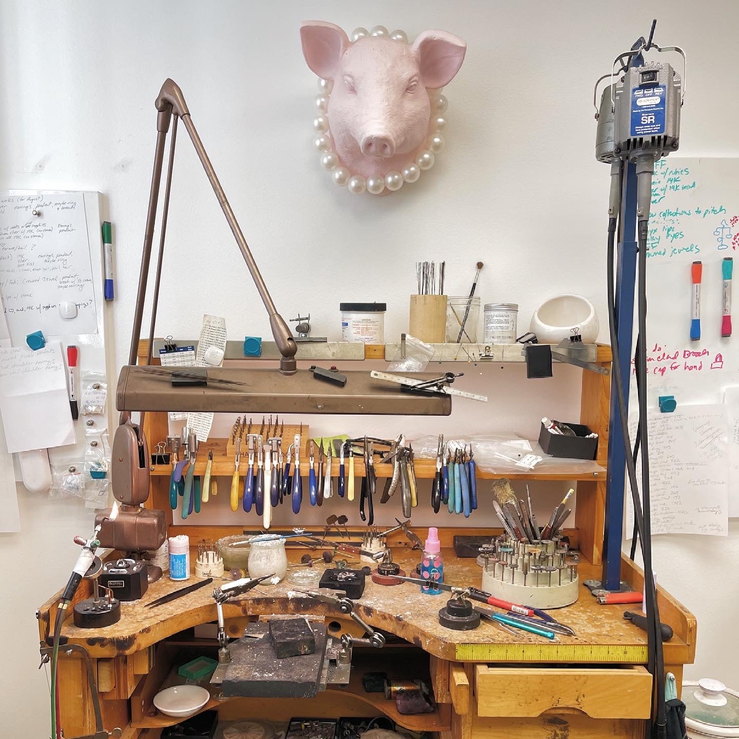Photo of a messy jeweler's brown wooden workbench with a pink pig head wearing pearls hanging on the white wall above it.
