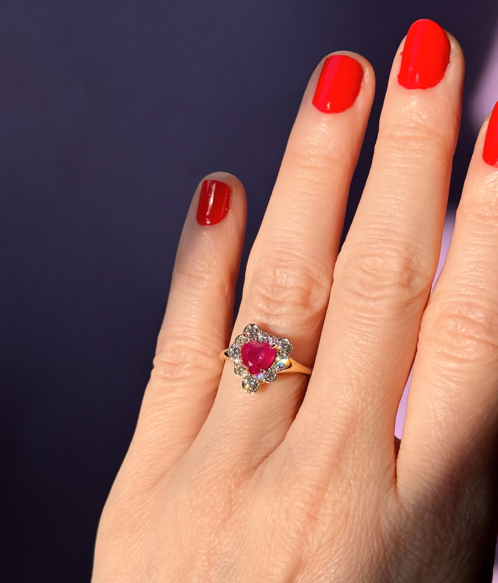 Hand wearing an 18K engagement ring with a ruby heart surrounded by diamonds against a dark background
