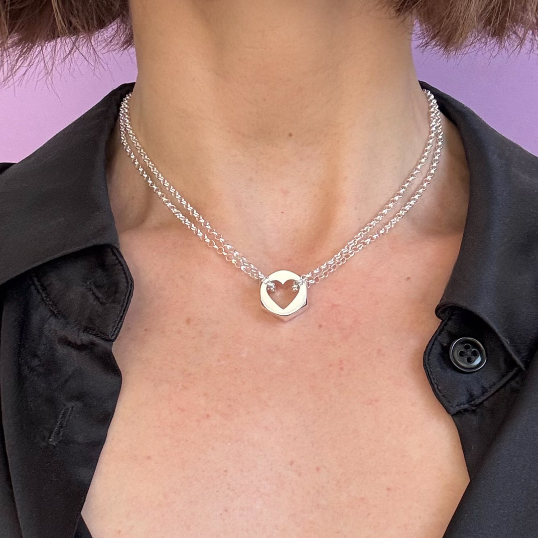 Person in a black shirt wearing a silver hex nut pendant on a silver chain with a heart-shape cut out of the center of the pendant, against a purple background