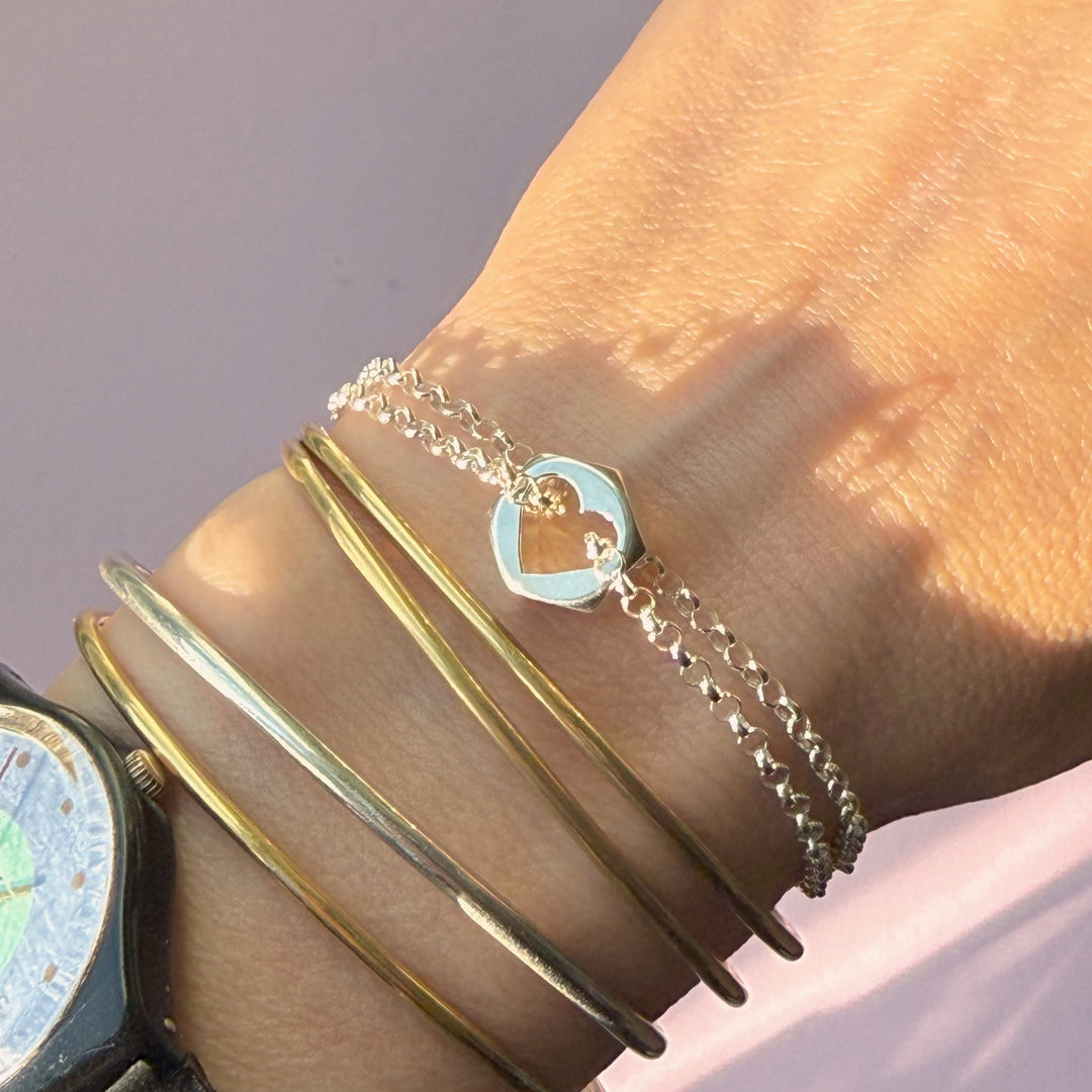Close-up of a wrist wearing gold bangles and a silver hex nut charm bracelet with a heart-shape cut out in the center, against a neutral background.