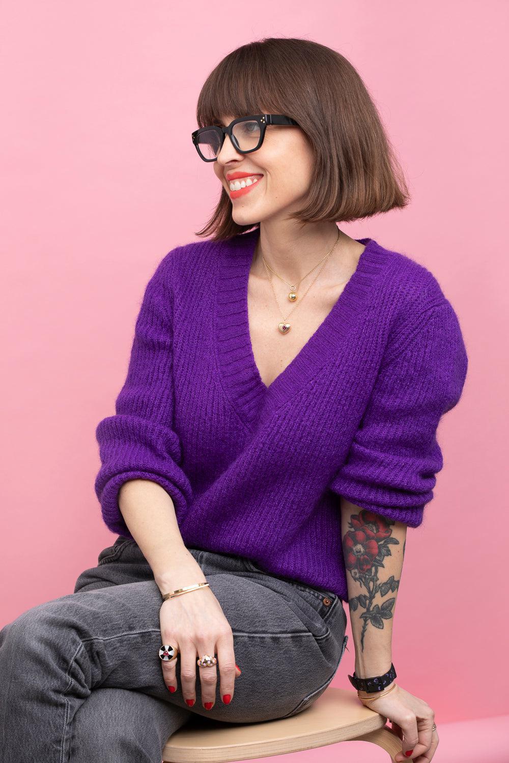Portrait shot of woman in purple sweater and grey jeans sitting on a stool against a pink background