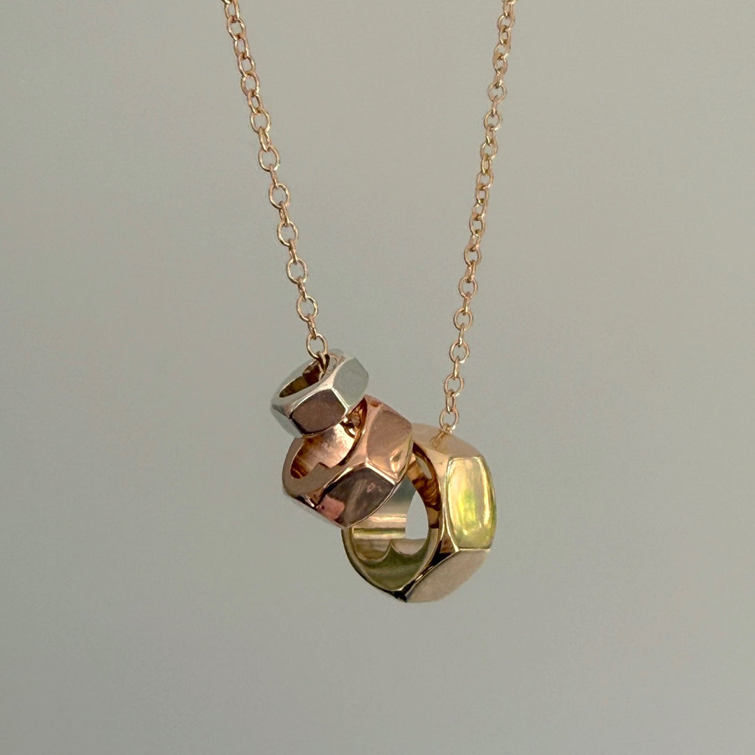 Close up of a gold chain with three hex nut pendants in silver, yellow and rose gold on a grey background.