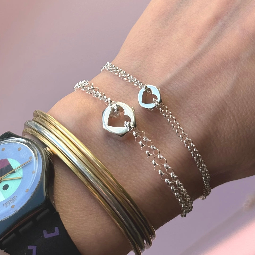 Wrist with multiple bracelets including two hex nut charms in silver with heart-shaped cuts in the center, against a neutral background