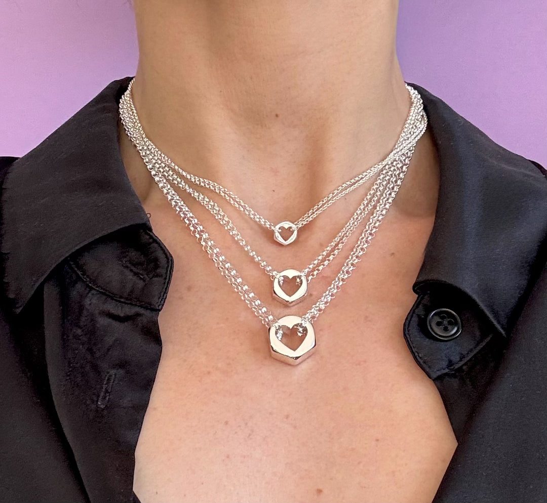 Person in a black shirt wearing three silver hex nut pendants on silver chains with heart-shapes cut out of the middle of each pendant, against a purple background