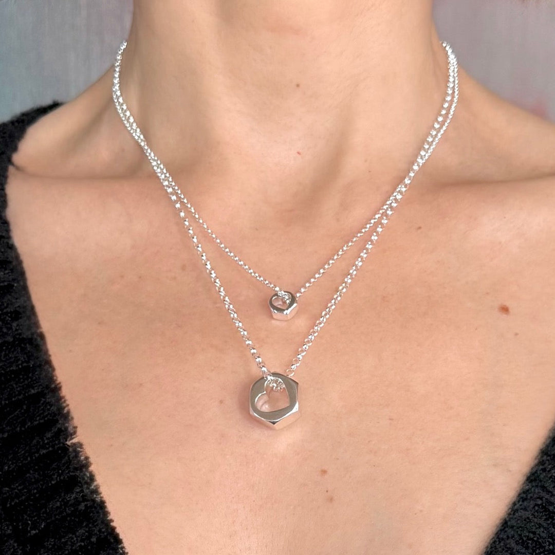 Close-up of a person wearing two silver hex nut pendant necklaces with heart cut outs in centers against a grey background.
