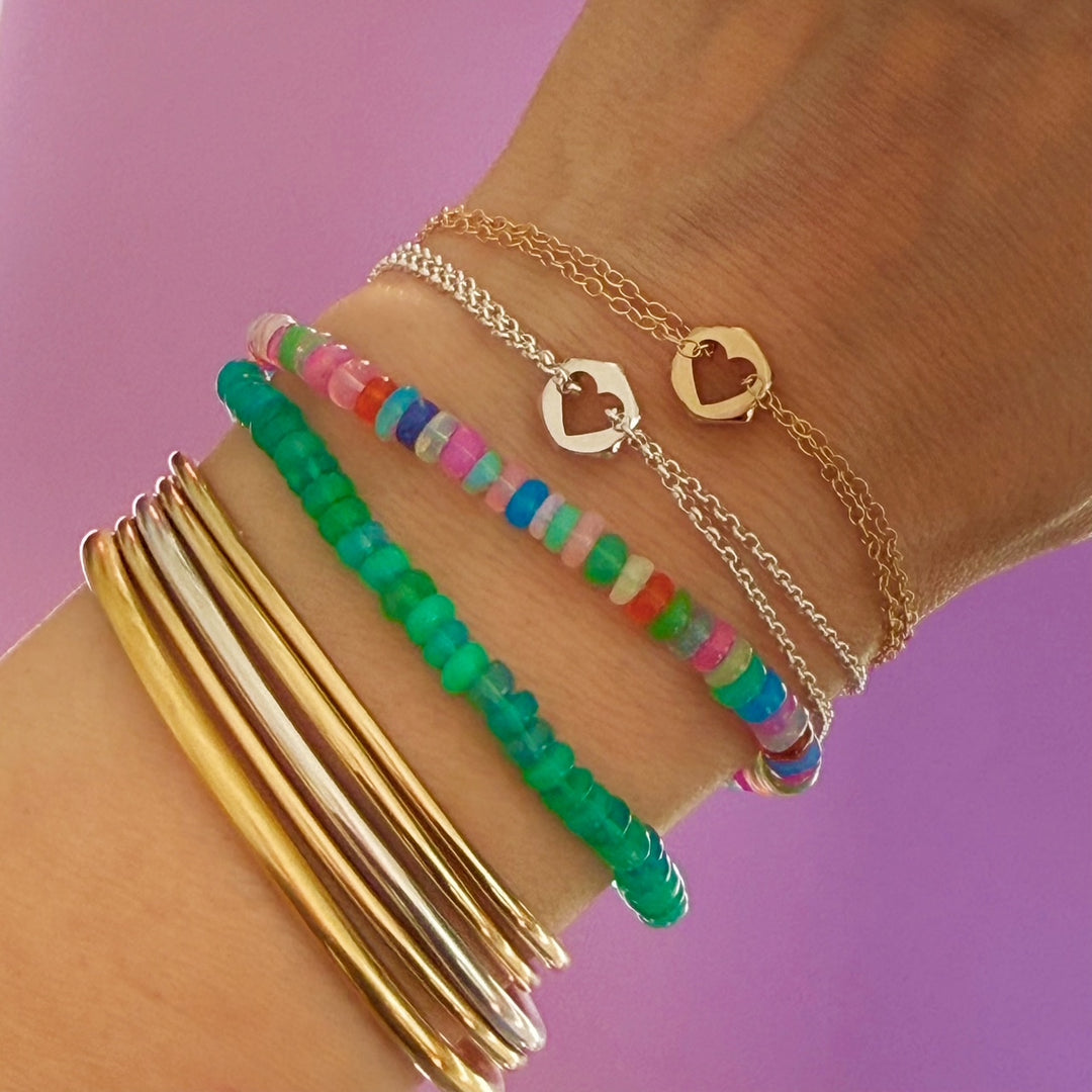 Close up of a wrist with multiple gold a silver bangle bracelets, green and multi-colored bead bracelets, and two hex nut charm bracelets with heart cut outs in gold and silver against a purple background