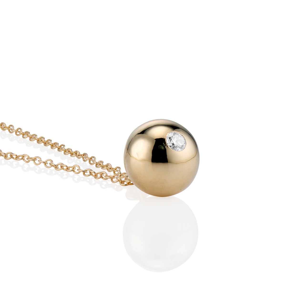 A gold ball pendant necklace with a small diamond set in bottom of ball, shown on a white background with a soft reflection.