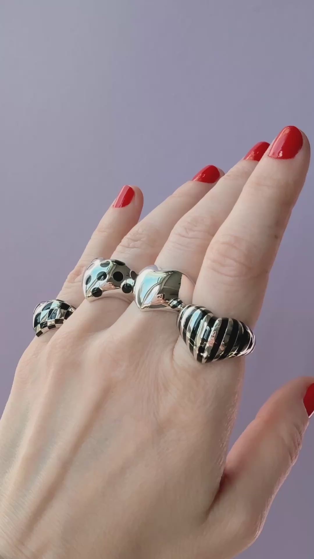 Video of a hand with red nails wearing four silver heart rings with various black patterns against a light purple background