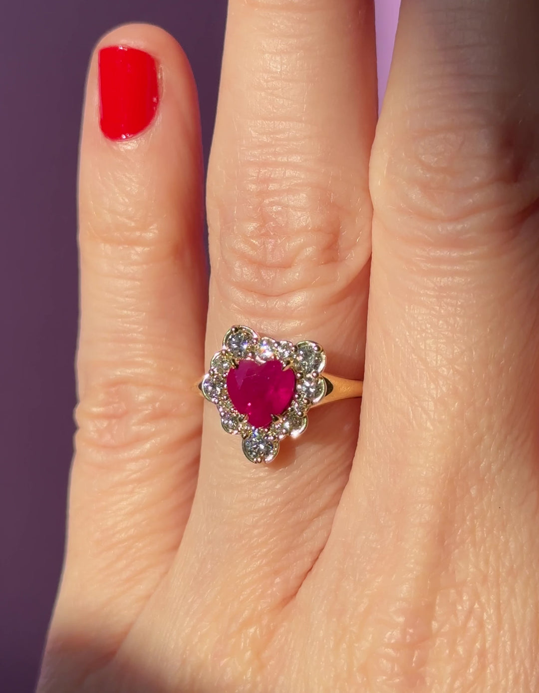 Video of a hand wearing an 18K engagement ring with a ruby heart surrounded by diamonds against a dark background