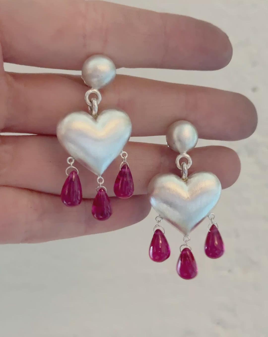 Video of silver heart earrings with ruby droplets dangling from below held between fingers against a white background