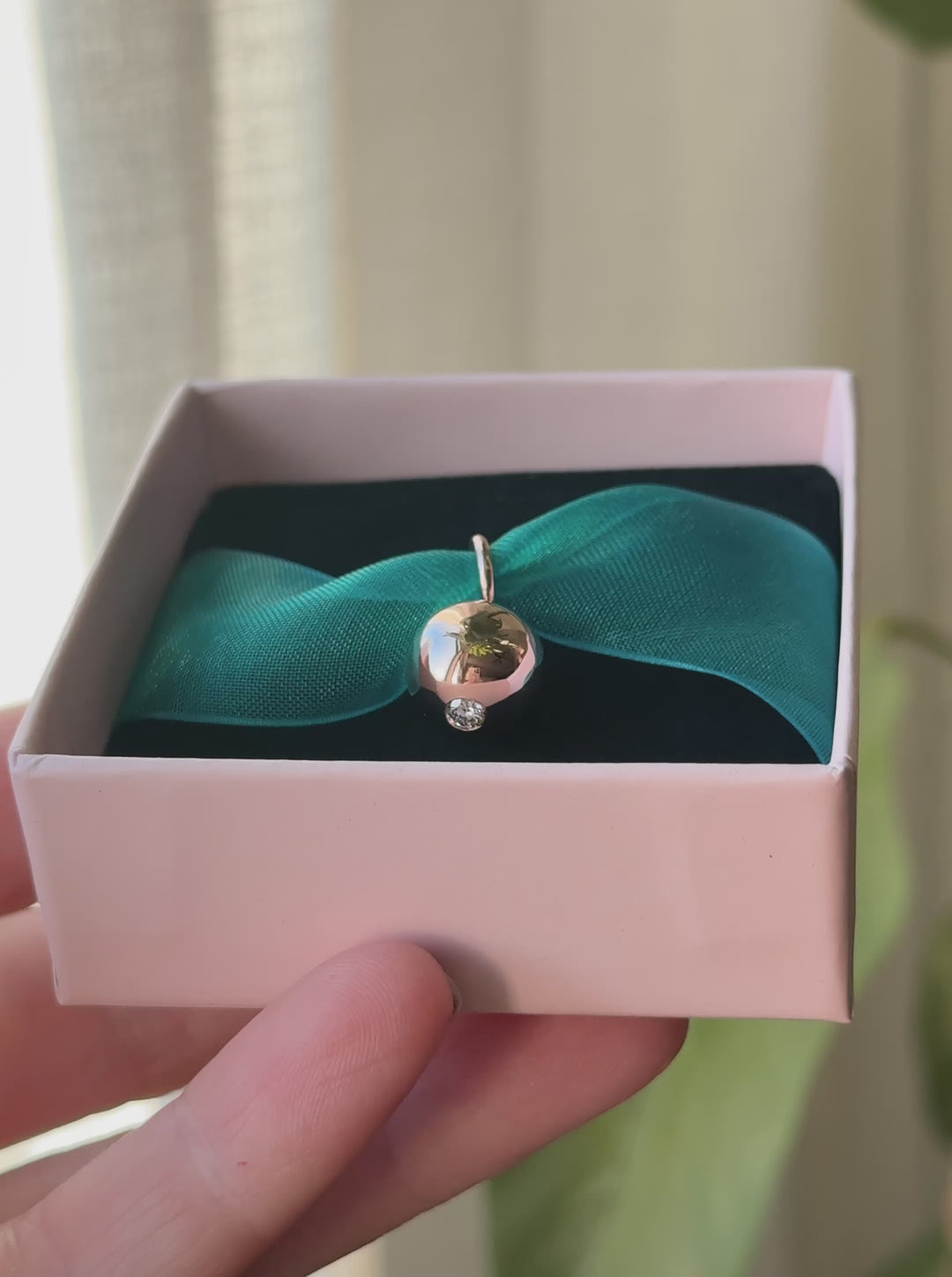 Video of a round rose gold charm with a diamond set into the bottom strung on a teal ribbon within a pink and black gift box.
