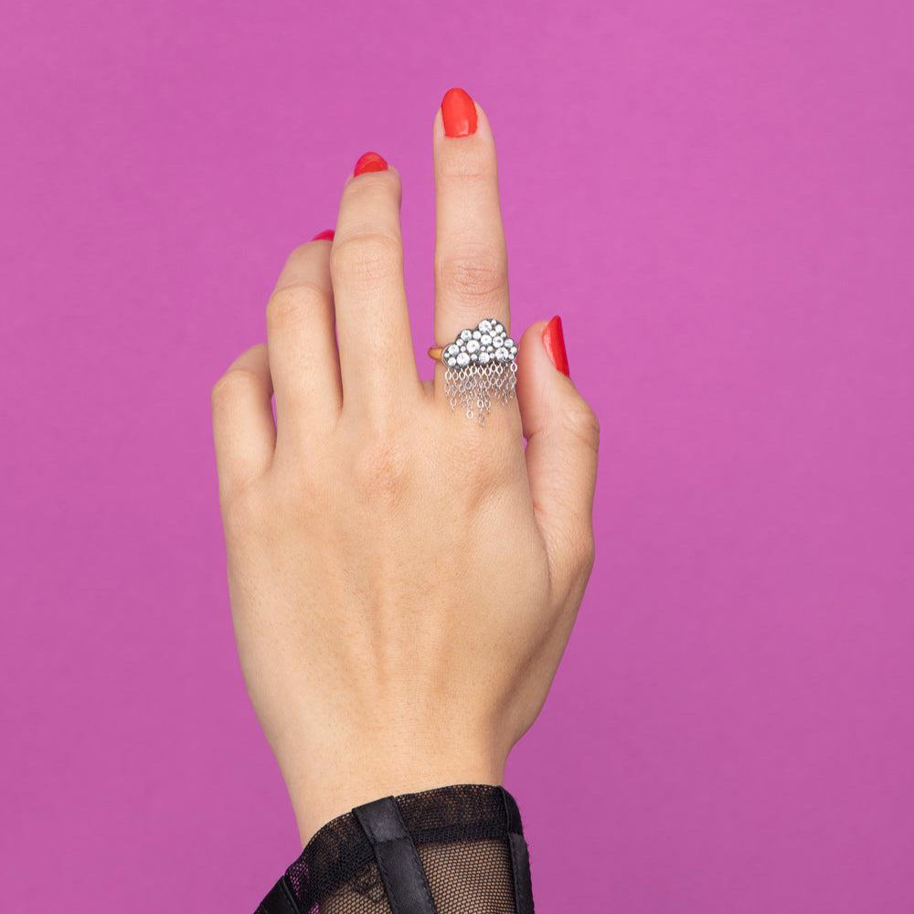 Rachel Quinn Jewelry black cloud shaped ring with pave white sapphires on cloud and dangling chains on yellow gold band on female model hand with purple background.