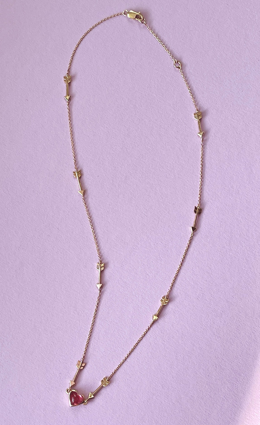 Yellow gold chain on a light purple background with 8 small arrows spaced intermittently, all pointing to the pink heart at the bottom center of the necklace
