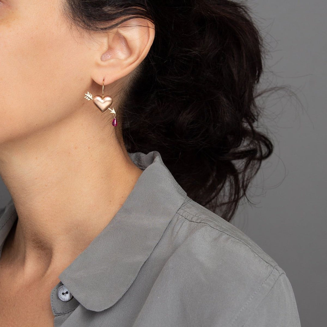 woman in grey blouse modeling puff heart hook earring in rose gold with yellow gold arrow piercing heart with lab grown ruby dripping from tip of arrow
