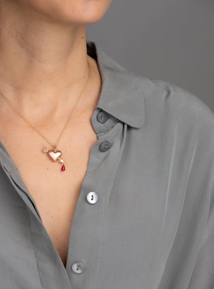 woman in grey blouse modeling small rose gold puffed heart pendant on yellow gold chain with yellow gold arrow piercing the heart with a single ruby dripping from tip of arrow