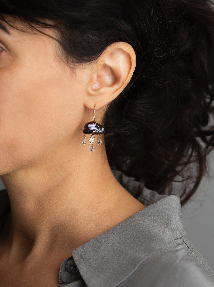 woman in grey modeling a single dark stormy keshi cloud like pearl earring with 14K yellow gold lightning bolt as a trio of white topaz rain down with gold ear wire