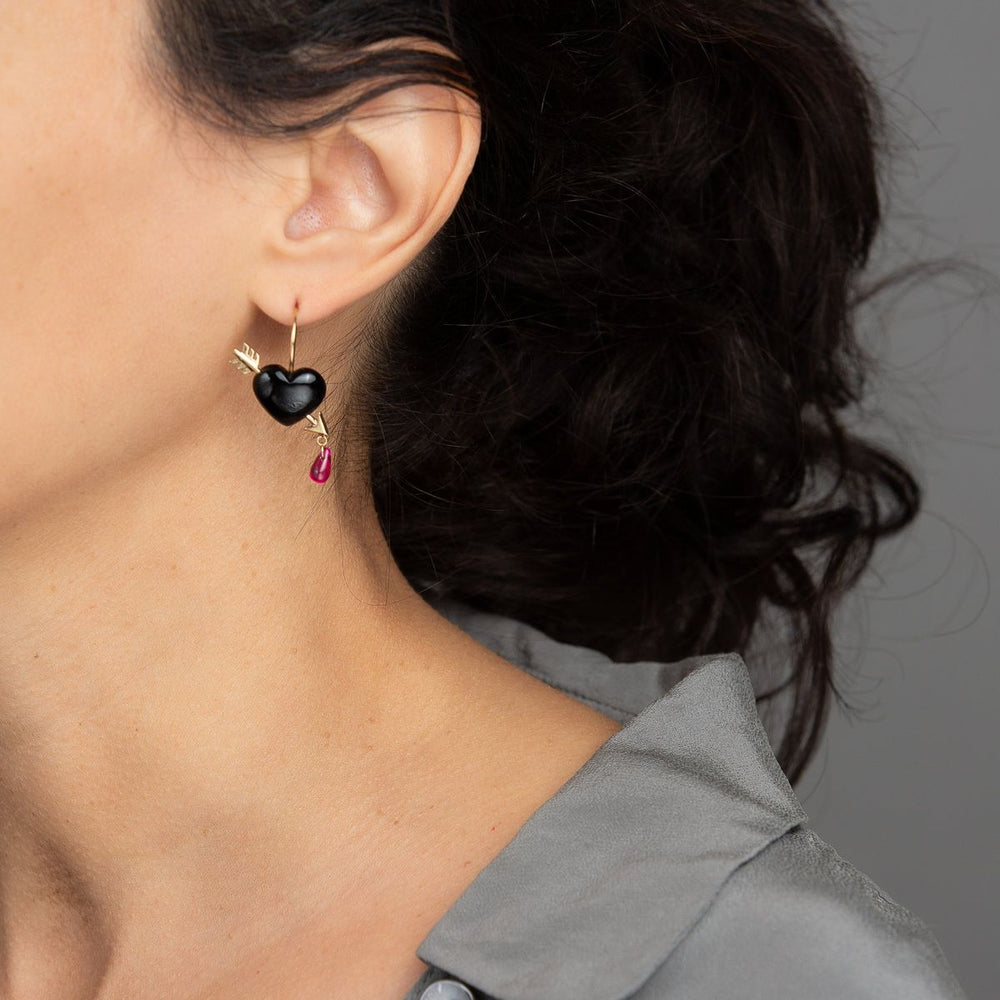 woman in grey models a single petite smooth jet black onyx heart earring is pierced with 14K gold arrows, leaving a single drop of ruby blood dripping from its tip