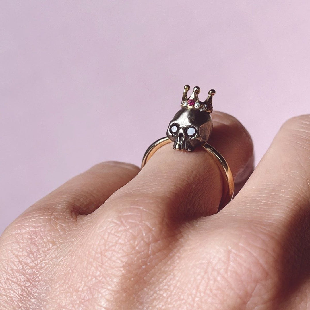 gold ring with blackened royal skull head with gold tooth adorning a ruby and diamond crown on model's ring ringer on pale pink background