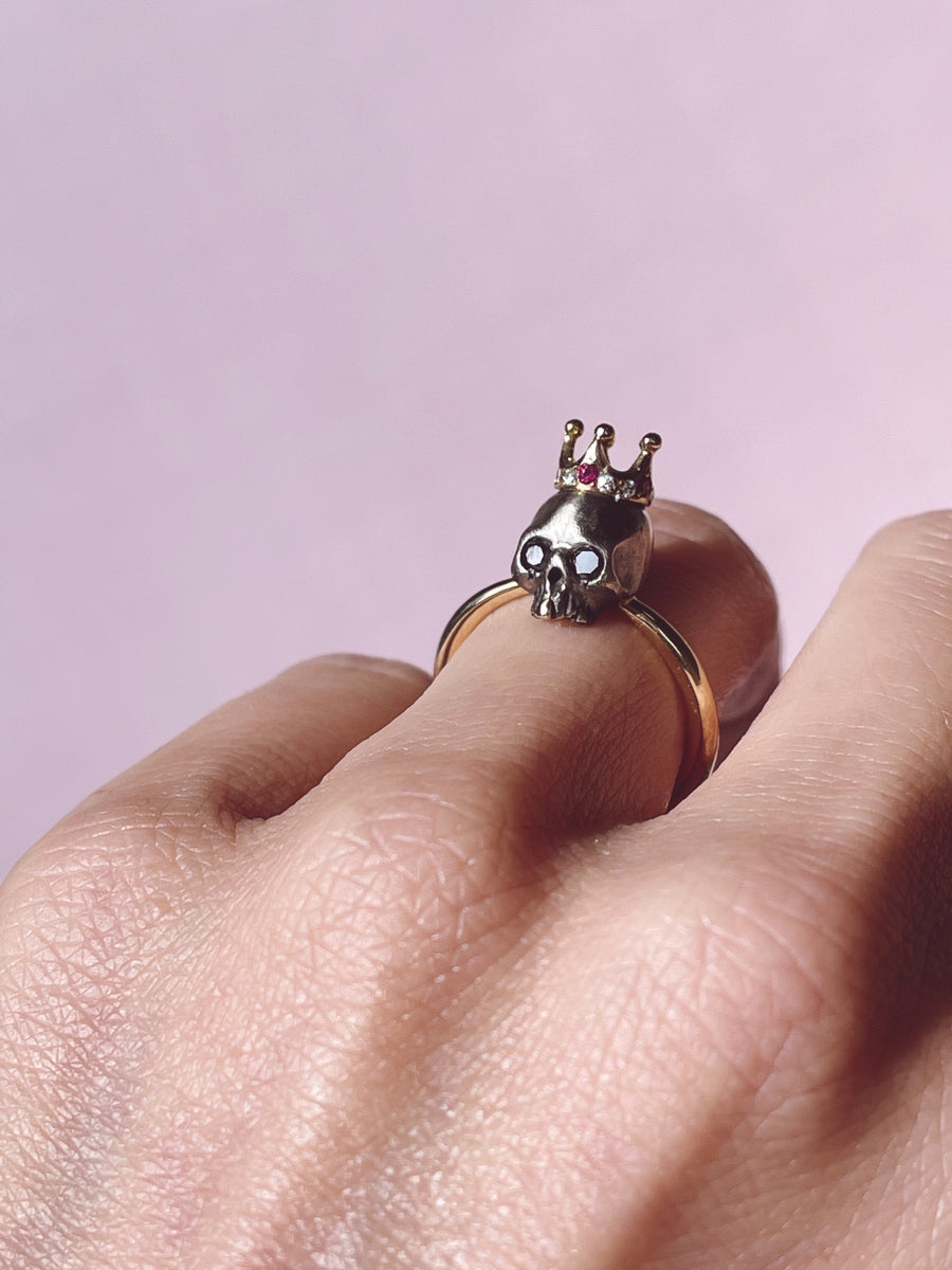 gold ring with blackened royal skull head with gold tooth adorning a ruby and diamond crown on model's ring ringer on pale pink background