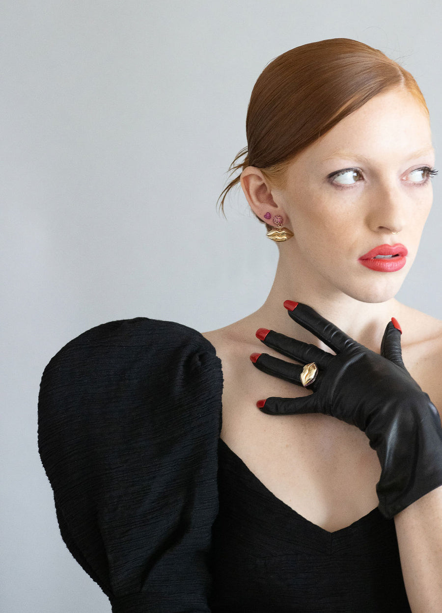 red head model wearing gold lips earrings and gold lip ring on her hand over black gloves next to her mouth on a grey background
